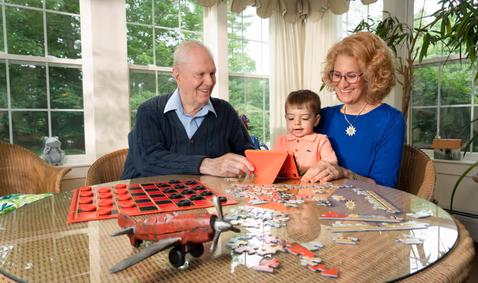 An elderly man and woman sitting at a round glass table with a young boy. They are engaged with a tablet and surrounded by puzzle pieces and a checkers board. The room has large windows with a view of green trees outside.
