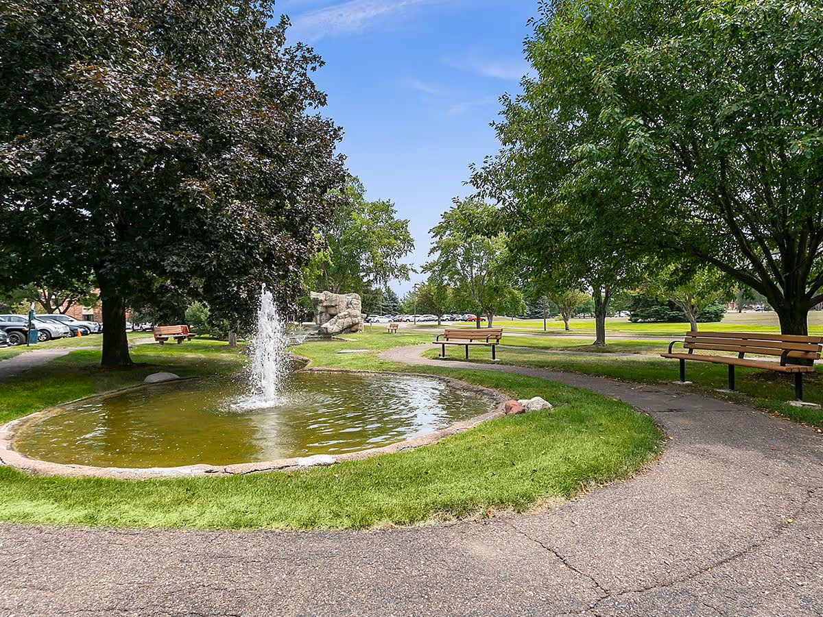 A peaceful outdoor park area with a small circular pond featuring a water fountain in the center. Surrounding the pond are green grassy areas, several trees providing shade, and multiple wooden benches along a curved paved pathway. Cars are visible parked in the background under a clear blue sky.