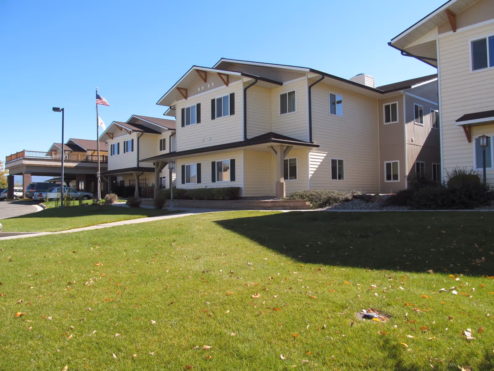 Exterior view of a two-story senior living facility building with beige siding and brown roof accents, surrounded by a well-maintained green lawn under a clear blue sky. There are two flagpoles with flags near the entrance and a covered parking area with cars visible.