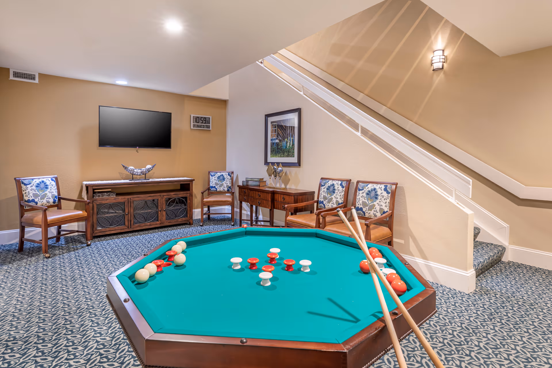 A cozy game room with a green octagonal billiards table in the center, featuring cue sticks and billiard balls. The room has patterned carpet flooring, beige walls, and a staircase with white railing. There are four wooden chairs with floral cushions arranged around the room, a wooden cabinet with a decorative piece on top, a wall-mounted flat-screen TV, and a framed picture on the wall.