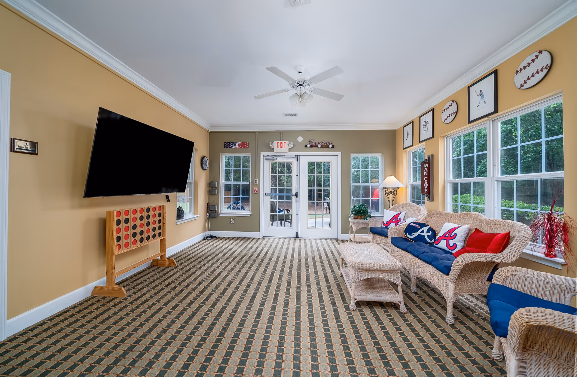 A bright and spacious living room with large windows letting in natural light. The room features wicker furniture with blue cushions and pillows decorated with the Atlanta Braves logo. A large flat-screen TV is mounted on a beige wall above a wooden game board. The walls are adorned with baseball-themed artwork and decorations. The room has a patterned carpet and a ceiling fan, with double glass doors leading outside.