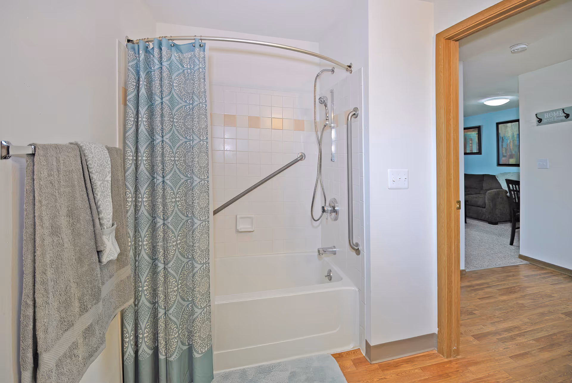 Bathroom with a bathtub and shower (curved rod and patterned curtain), grab bars, a towel rack, and an open door into a living area.