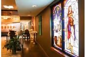 Interior hallway of a senior living facility with stained glass windows on the right side and a person walking in the distance. The hallway has wooden flooring, plants, and soft lighting.