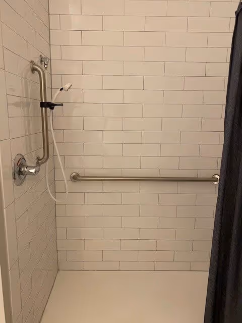 A tiled shower area with white rectangular tiles, featuring a handheld showerhead attached to a vertical grab bar and a horizontal grab bar on the wall. A dark shower curtain is partially visible on the right side.