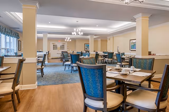 A spacious dining room with multiple tables and chairs arranged neatly. The chairs have blue patterned backs and cream-colored seats. The room features light yellow walls, white trim, and a combination of wood and carpeted flooring. Chandeliers hang from the ceiling, and framed artwork is visible on the walls.