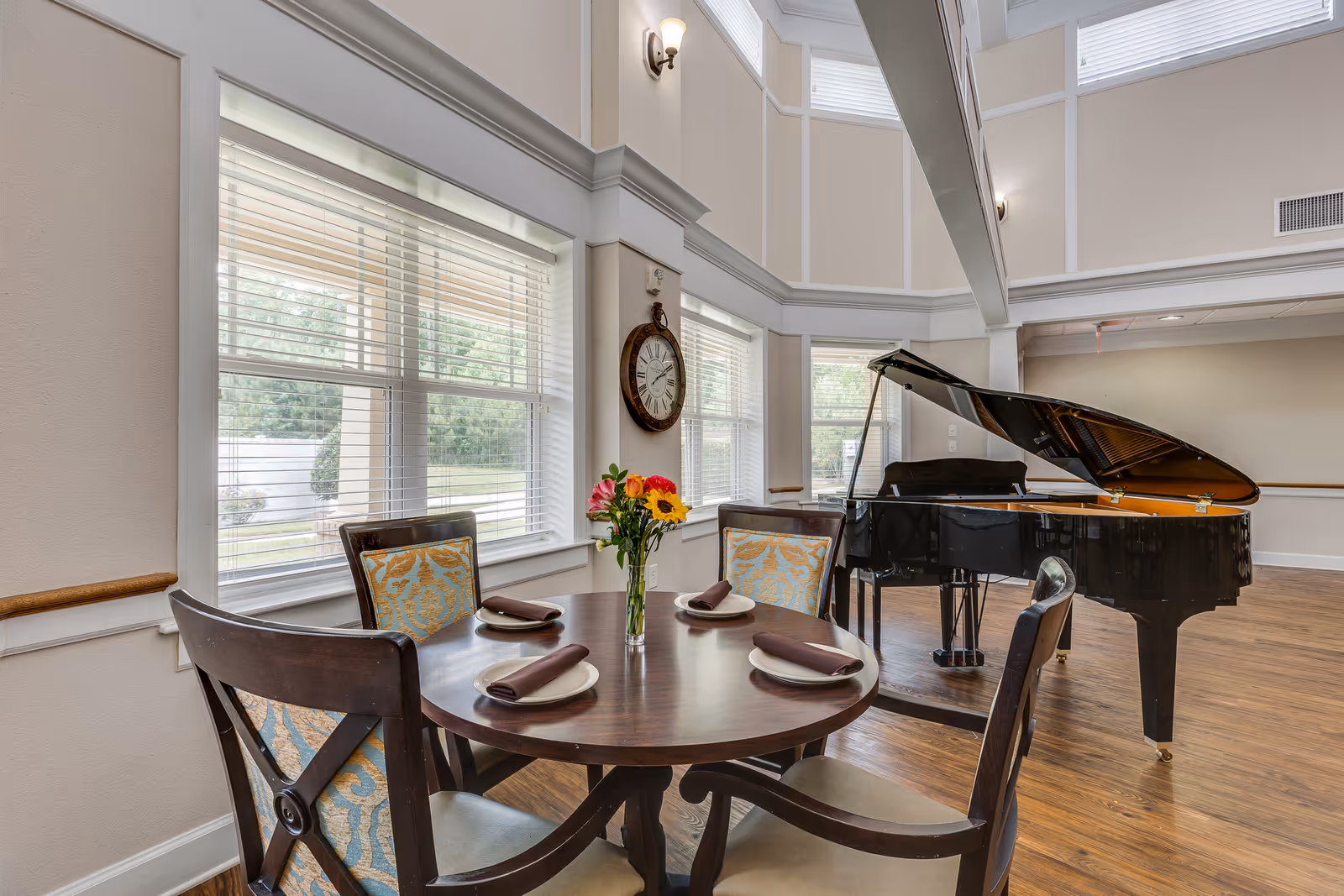 A bright communal dining area with a round table set for four and a black grand piano by tall windows.