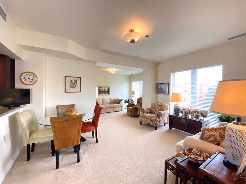 A bright and spacious living room area with beige carpeting and white walls. The room features a glass dining table with four chairs in different colors, a beige sofa, a floral armchair, and a recliner. There are two large windows with blinds letting in natural light. The room is decorated with framed artwork, table lamps, and various small decorative items on tables and a wooden chest.
