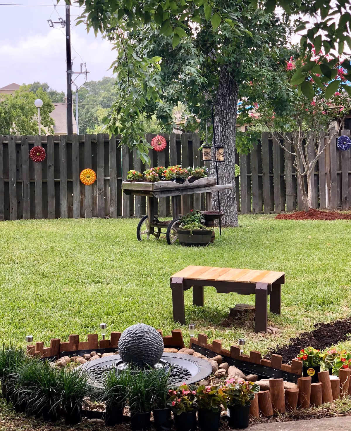 A fenced backyard garden featuring a small spherical water fountain surrounded by plants, a wooden bench, potted flowers, and a vintage planter cart under a tree.