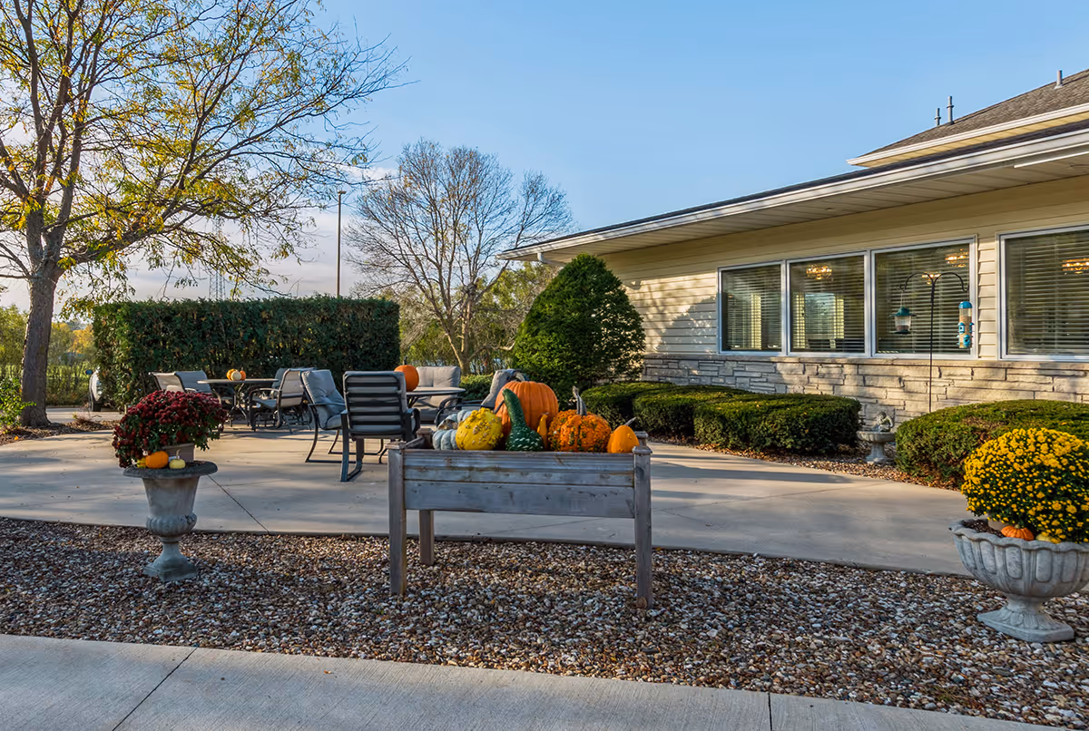 Outdoor patio area at Pinicon Senior Living with tables and chairs arranged for seating. The patio is decorated with pumpkins and gourds in a wooden planter, and there are potted flowers on either side. The building exterior features large windows and neatly trimmed bushes, with trees and a clear blue sky in the background.