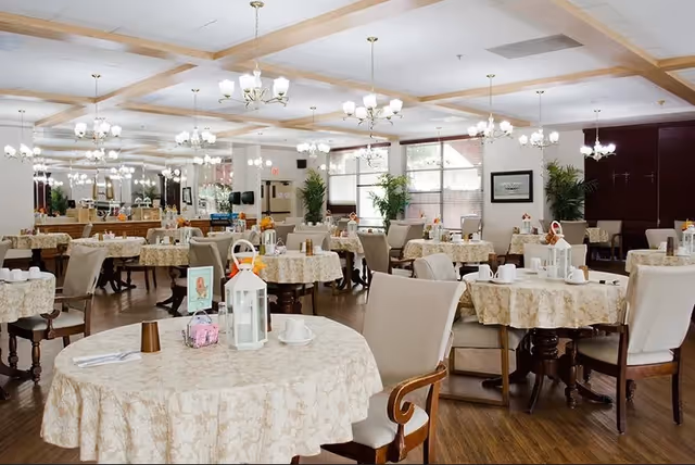 A spacious dining room with multiple round tables covered in beige patterned tablecloths. Each table is set with white cups, saucers, and decorative lanterns. The room features wooden flooring, beige cushioned chairs, large windows with blinds, and several chandeliers hanging from a coffered ceiling. There are plants and framed artwork on the walls, creating a warm and inviting atmosphere.