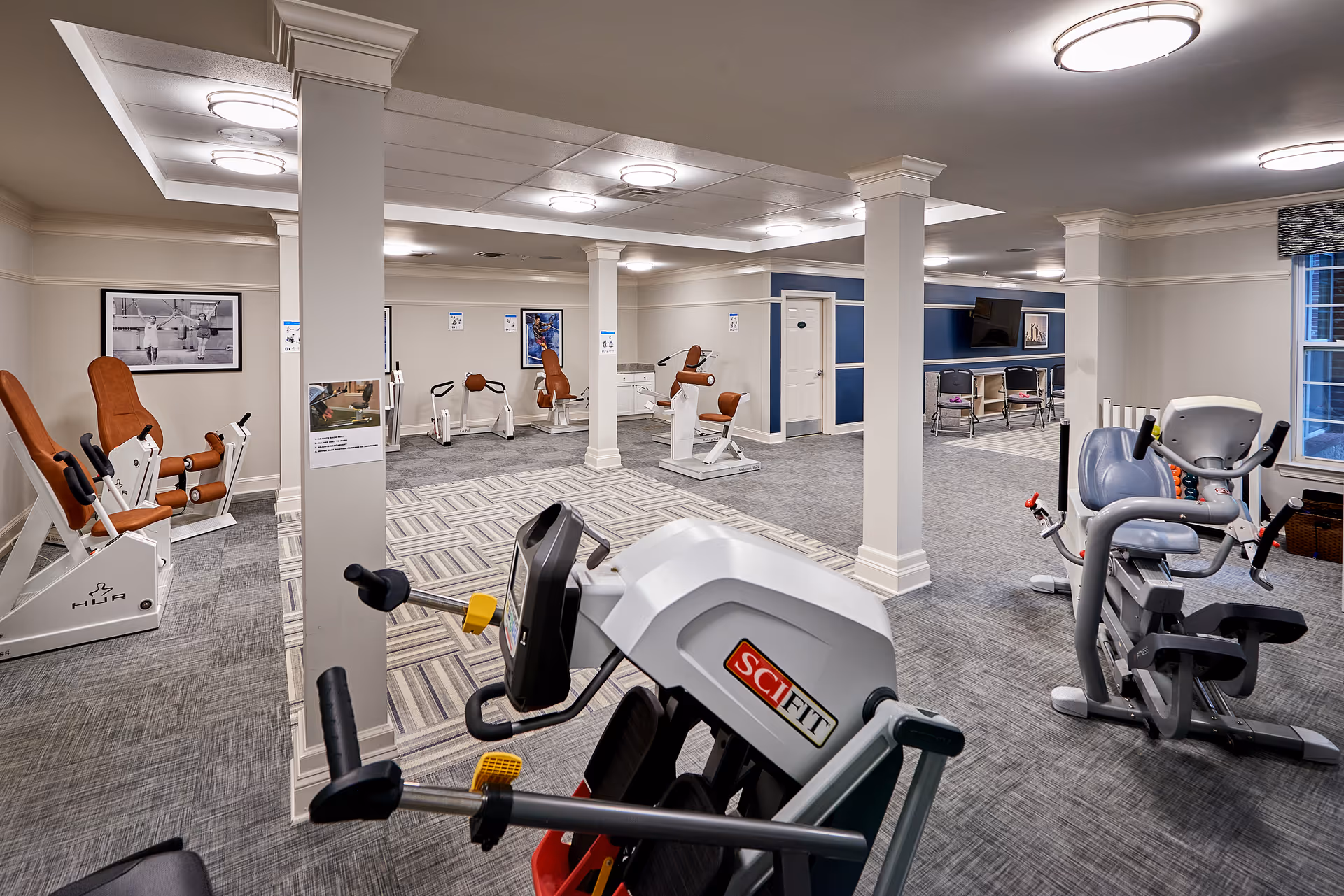A spacious fitness room in a senior living facility featuring various exercise machines with brown and gray seats, including a SciFit machine in the foreground. The room has gray carpeted floors, white walls with blue accent panels, multiple white columns, and ceiling lights. There are framed pictures on the walls and a TV mounted on the far wall.