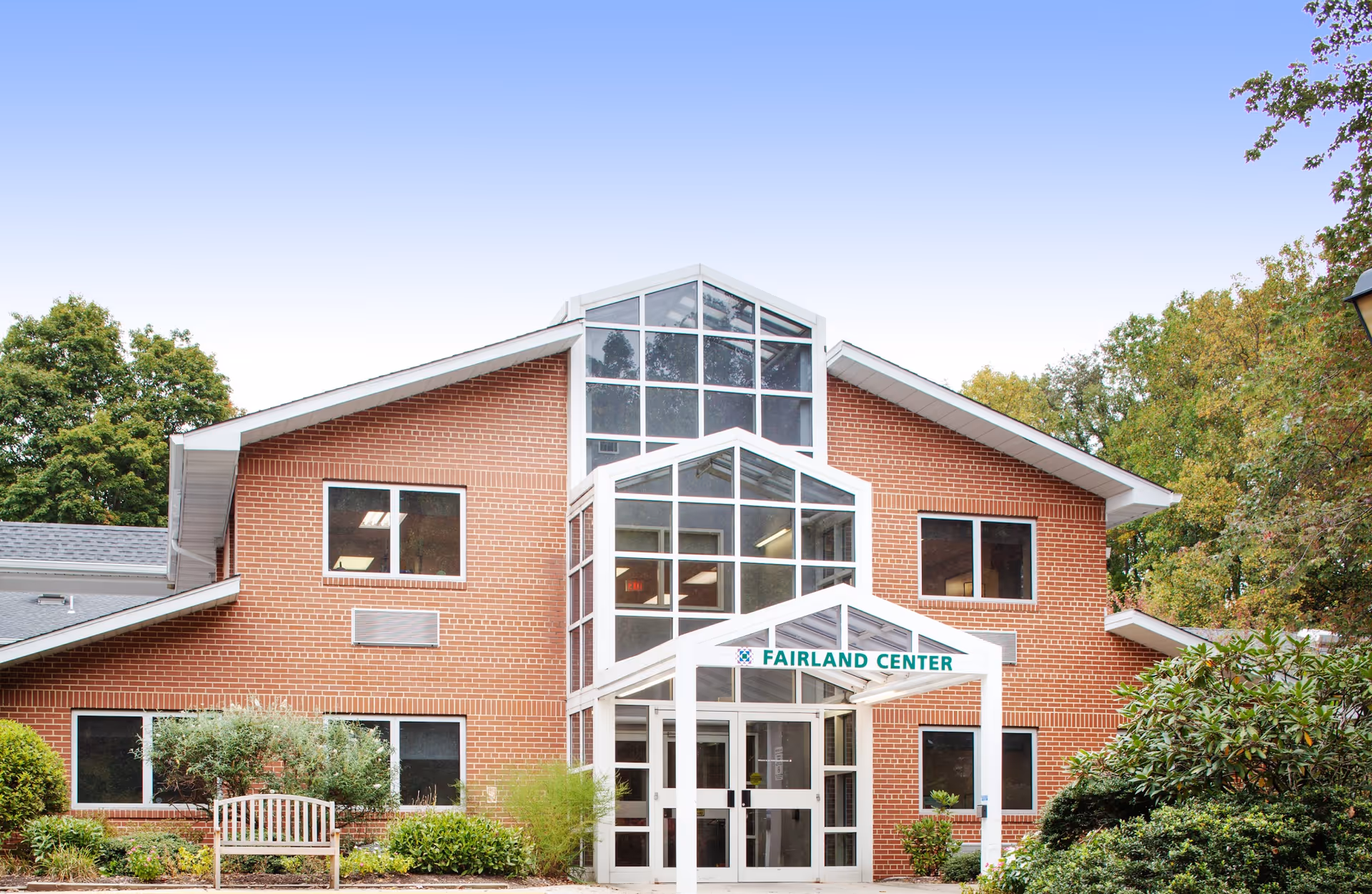 Exterior front view of Fairland Center, a two-story brick building with large glass windows and a white entrance canopy. The building is surrounded by green shrubs, trees, and a wooden bench in front.