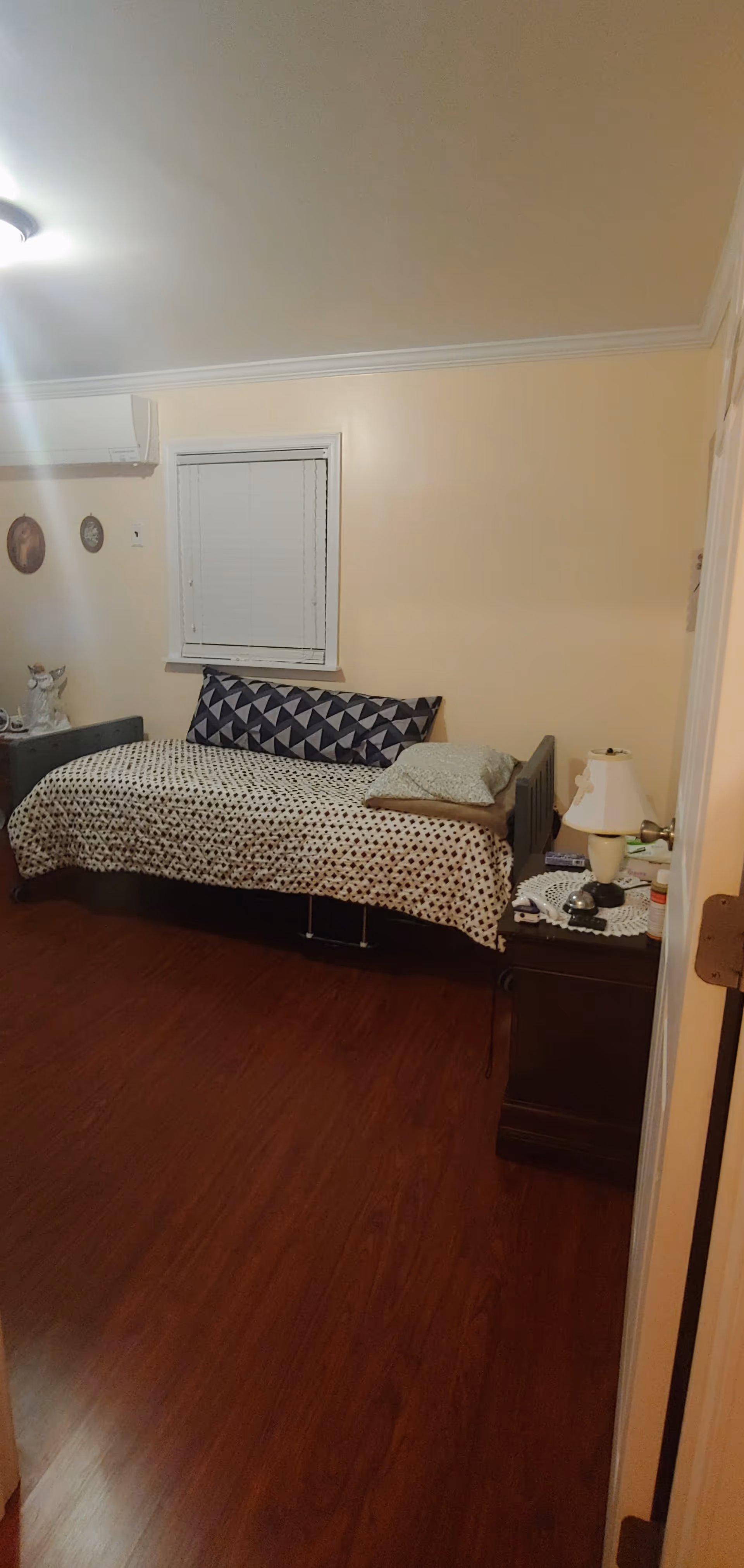A small bedroom with a single bed covered with a patterned blanket and pillows. There is a wooden nightstand next to the bed with a lamp, a bell, and some small items on a doily. The room has light-colored walls, a window with closed blinds, and wooden flooring. A wall-mounted air conditioning unit is visible above the bed.