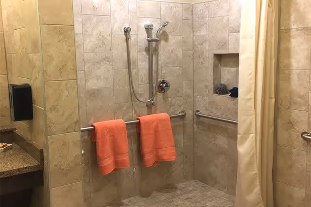 Accessible tiled walk-in shower with grab bars, a handheld showerhead, built-in niche, and two coral towels hanging on the bar.