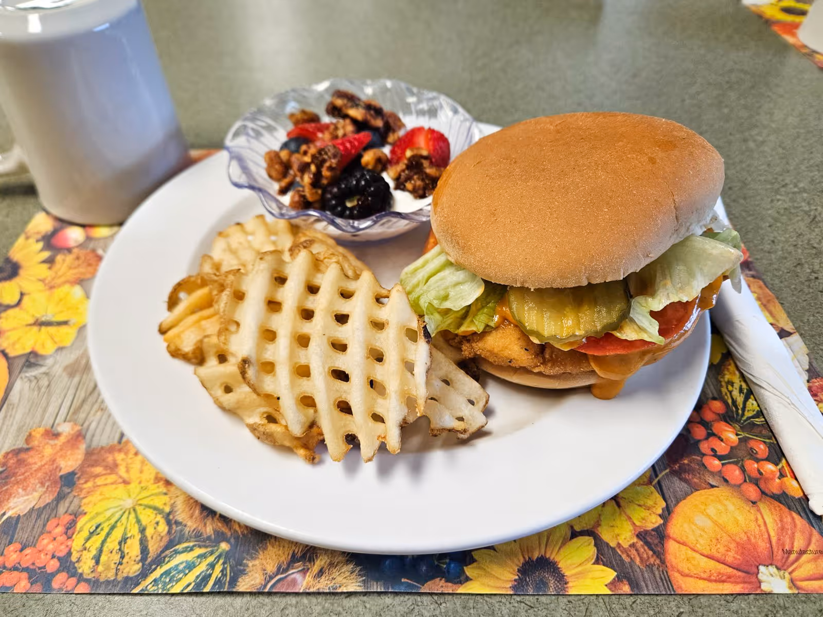 A plate with a chicken sandwich topped with pickles and lettuce, waffle-cut fries, and a small fruit-and-nut bowl on a floral placemat with a mug nearby.