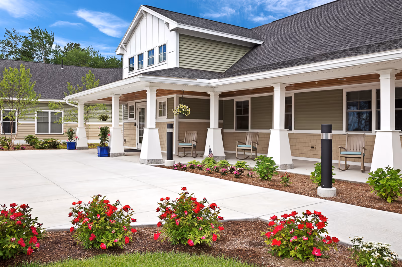 Front entrance of a senior living facility with a covered porch, columns, benches, and landscaped flower beds.
