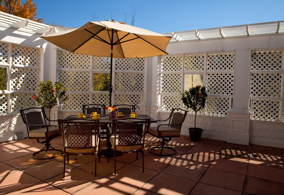Sunlit enclosed patio with a table, umbrella and chairs surrounded by white lattice walls and potted plants.