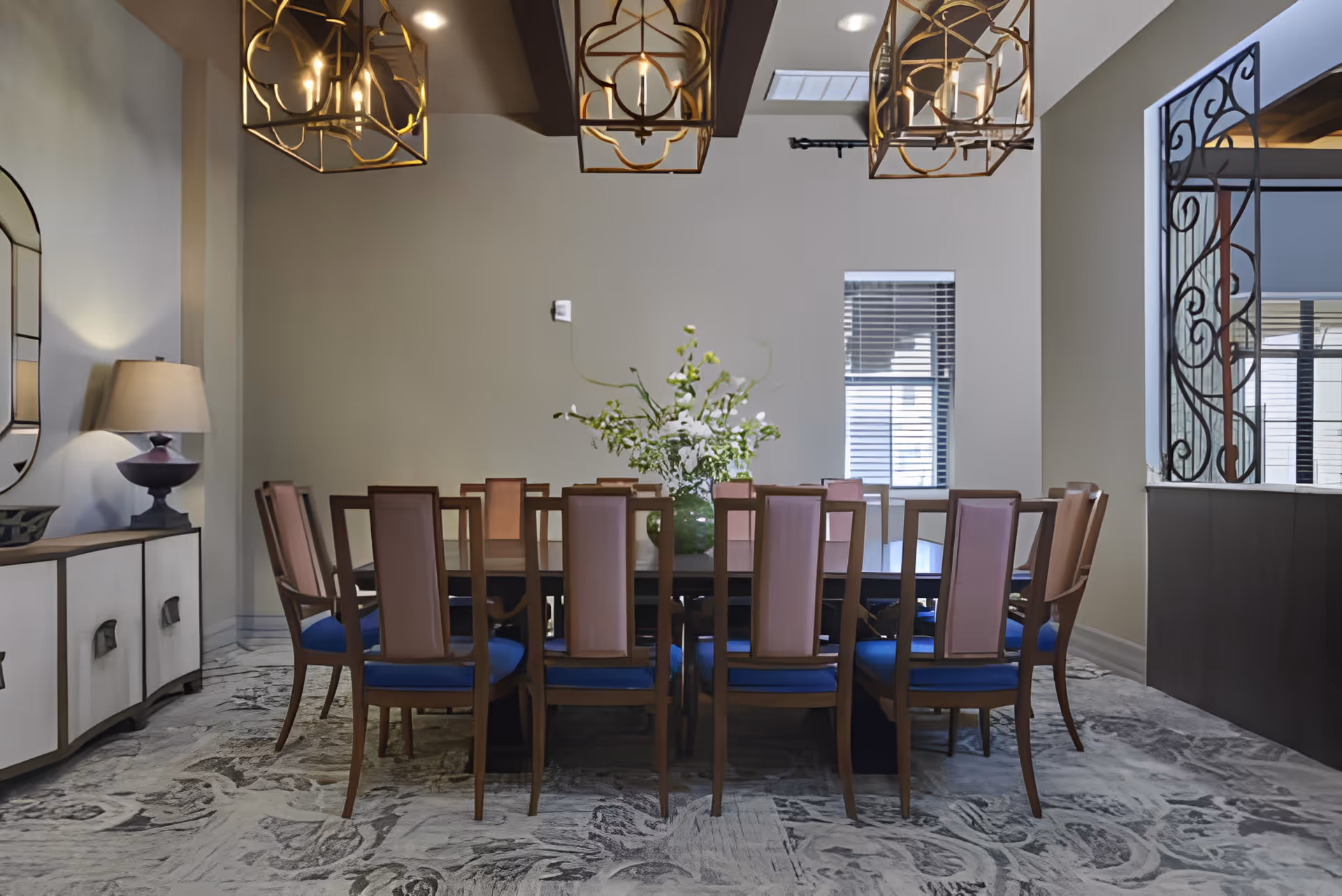 A formal dining room with a long rectangular table surrounded by wooden chairs with blue cushions and light pink backs. A large floral arrangement is centered on the table. The room features a patterned carpet, a sideboard with a lamp and mirror on the left wall, and three decorative gold pendant lights hanging from the ceiling. There is a window with blinds on the far wall and a decorative wrought iron partition on the right.