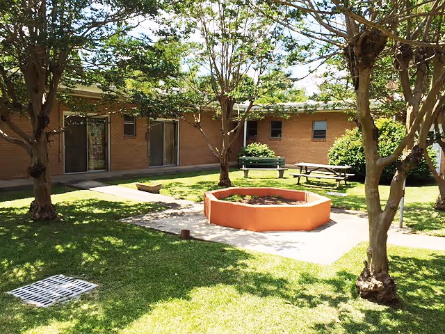 Outdoor courtyard area with green grass, several trees, a raised octagonal planter in the center, a green bench, and a picnic table. The courtyard is surrounded by a single-story brick building with windows and sliding glass doors.