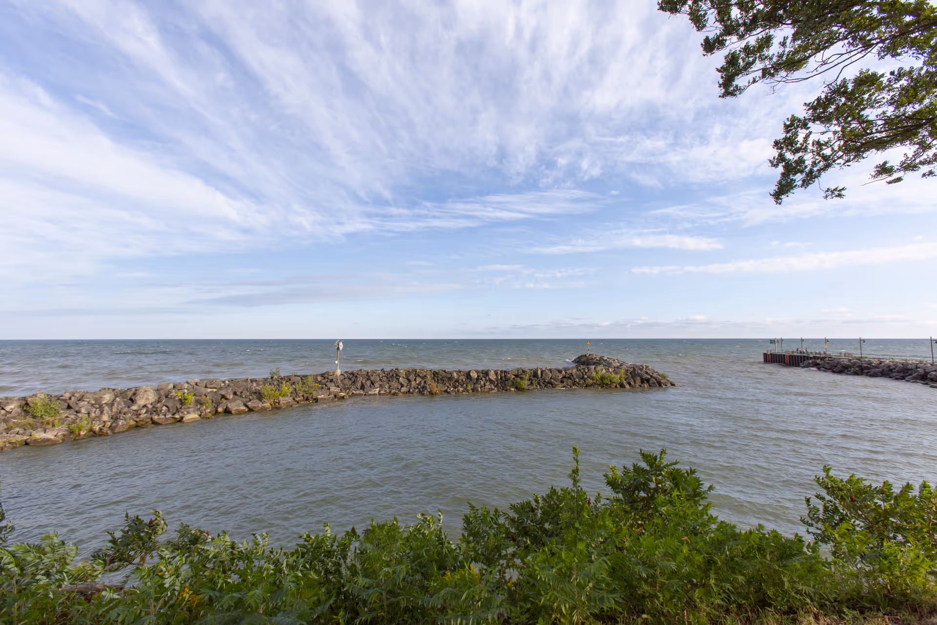 View of a calm body of water bordered by a rocky breakwater with some greenery in the foreground and a partly cloudy sky above.