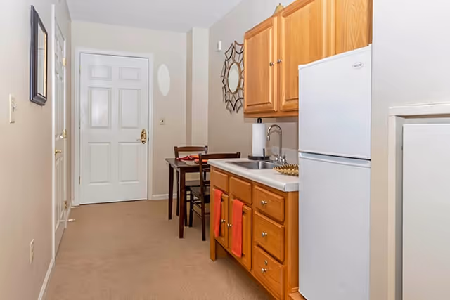 A small kitchenette area in a senior living facility with wooden cabinets, a white refrigerator, a sink, and a small dining table with two chairs against a beige wall. A decorative mirror hangs above the table, and the floor is carpeted.