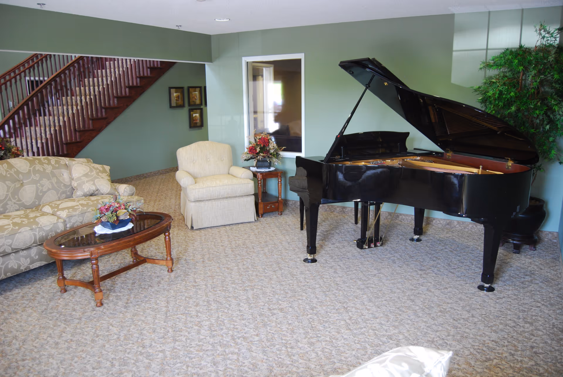 A senior living facility common area with a black grand piano, a beige armchair, a floral patterned sofa, a wooden coffee table with a flower arrangement, and a staircase with wooden railing in the background. The walls are painted light green and there is a large potted plant near the piano.