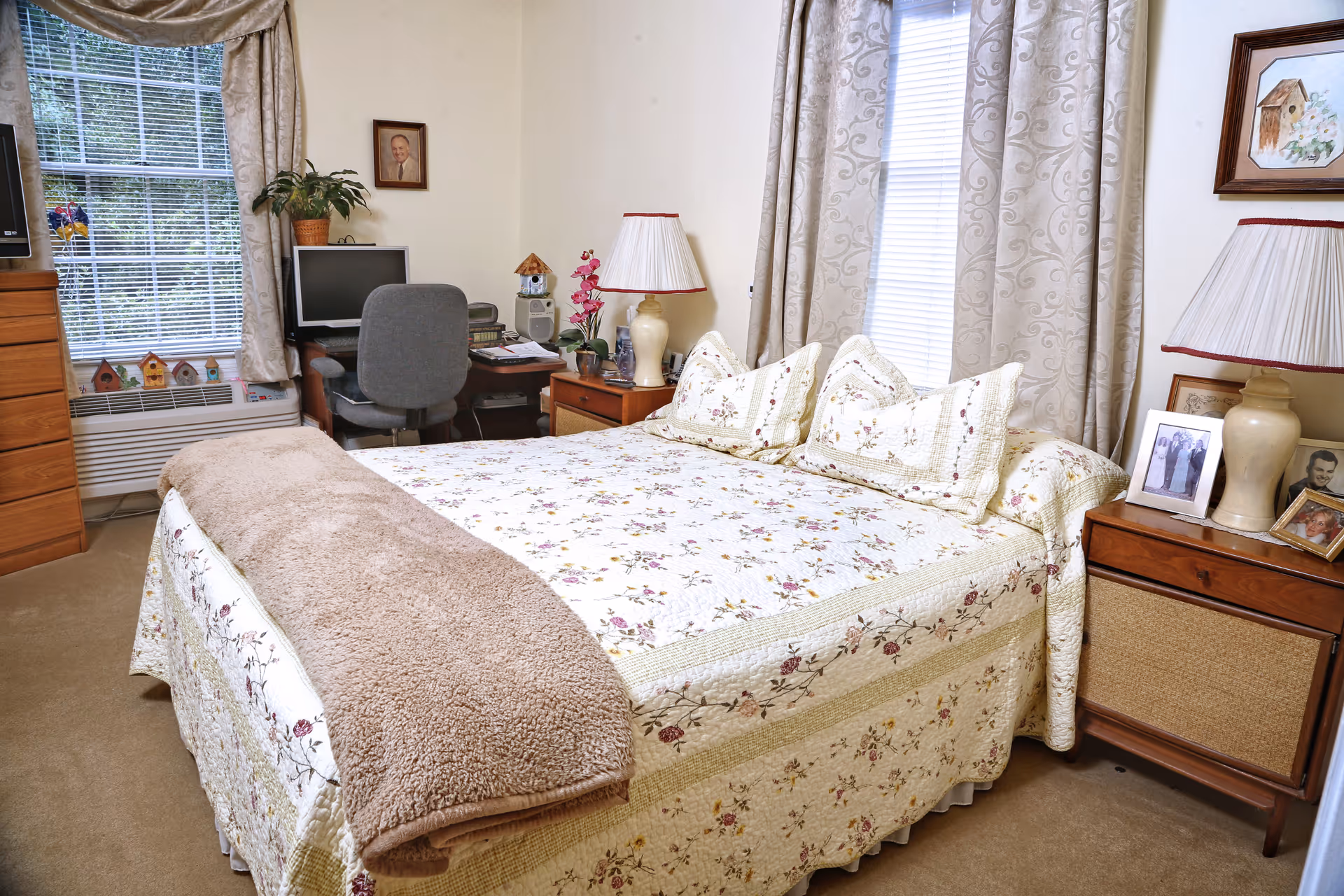 A cozy bedroom with a bed covered in a floral quilt and matching pillows. There is a beige blanket folded at the foot of the bed. On either side of the bed are wooden nightstands with lamps and framed photos. A window with patterned curtains is behind the bed. In the corner, there is a desk with a computer, a gray office chair, and some decorative items including a plant and small birdhouses on the windowsill.