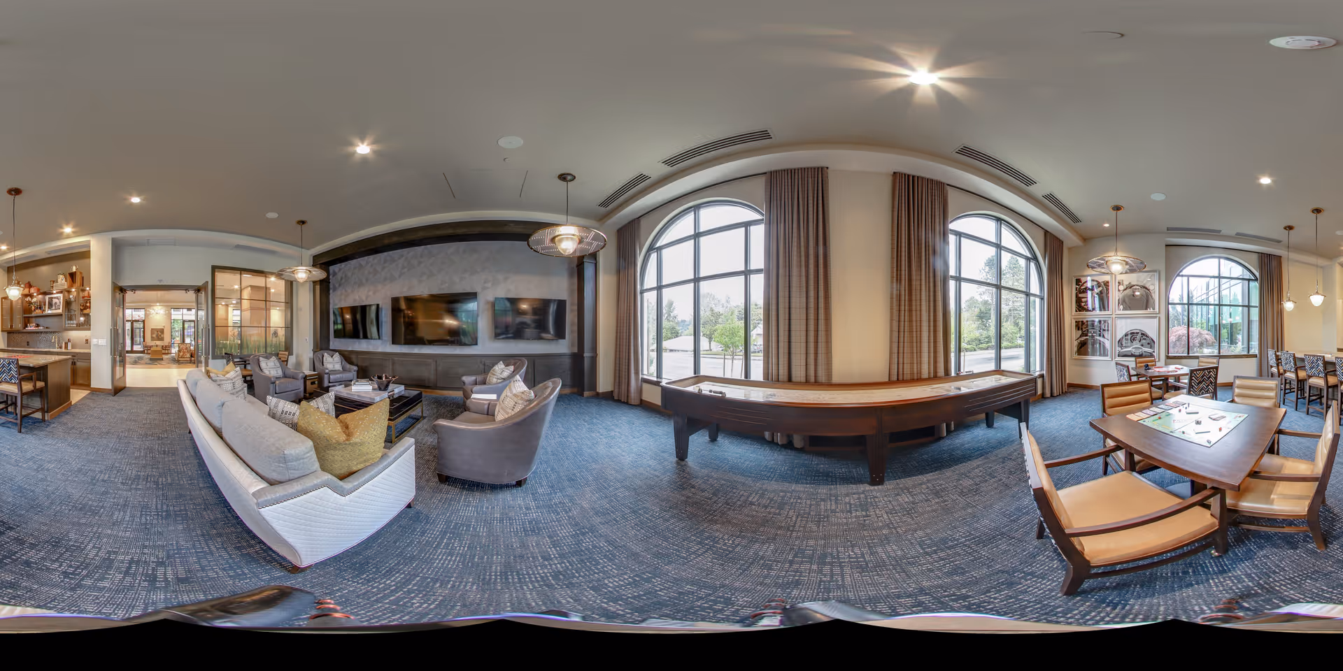 Bright communal lounge with seating around a wall of TVs, a shuffleboard table by arched windows, and a nearby dining/game area.