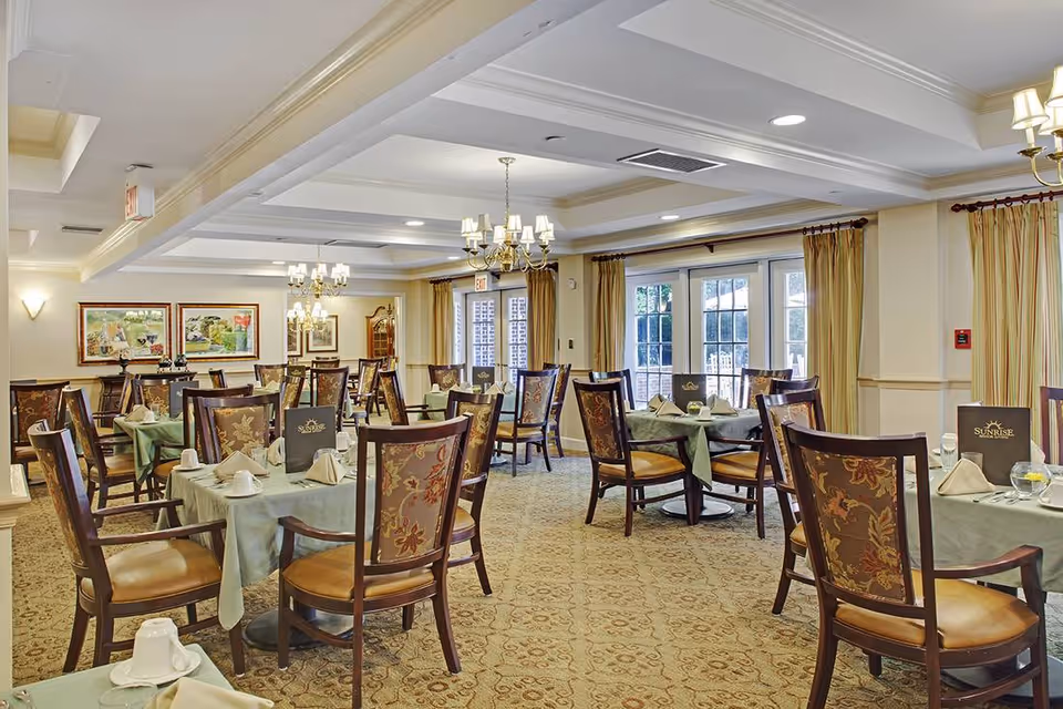 A spacious dining room with multiple tables covered in light green tablecloths, each set with white napkins, cups, and menus. The room features elegant wooden chairs with floral upholstery, chandeliers hanging from a coffered ceiling, large windows with beige curtains, and framed artwork on the walls.