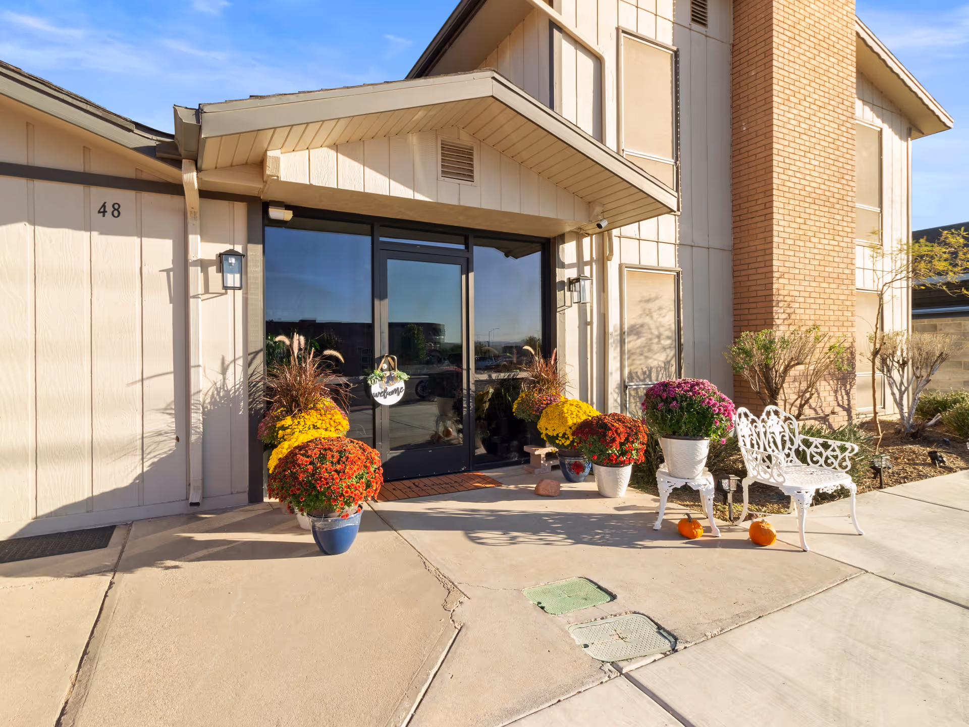Entrance of a building with glass double doors, decorated with colorful potted flowers and two small pumpkins on the ground near a white metal bench. The building has beige siding and a brick chimney, with the number 48 visible on the left side.