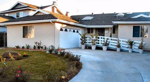 Single-story tan home with white porch railing, potted plants, and curved driveway.