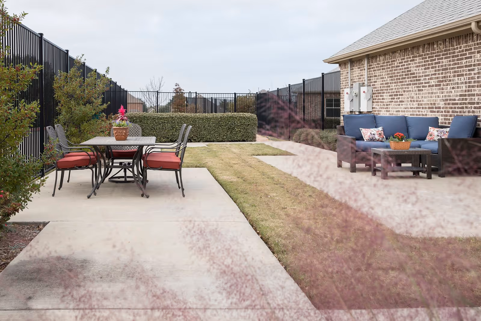 Outdoor patio area with a dining table and six chairs with red cushions on the left side, and a seating area with a blue cushioned sofa and chairs with floral pillows on the right side, adjacent to a brick building. The area is fenced with black metal fencing and has some bushes and grass.