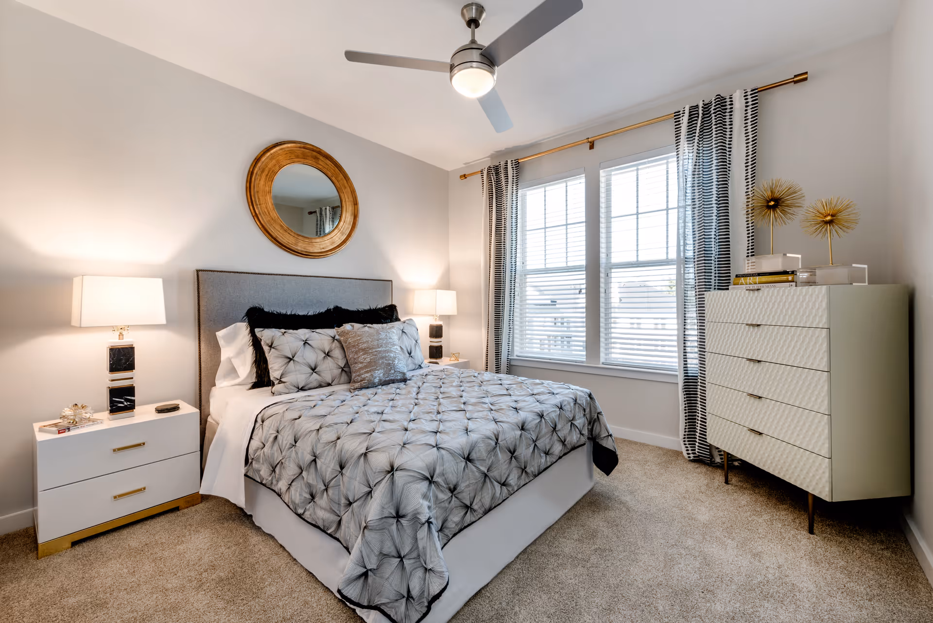 A neatly made bed with a tufted gray and black comforter and multiple pillows in a bright bedroom. The room features a round gold-framed mirror above the bed, two bedside tables with matching lamps, a ceiling fan with a light, a window with black and white striped curtains, and a white dresser with decorative items on top.