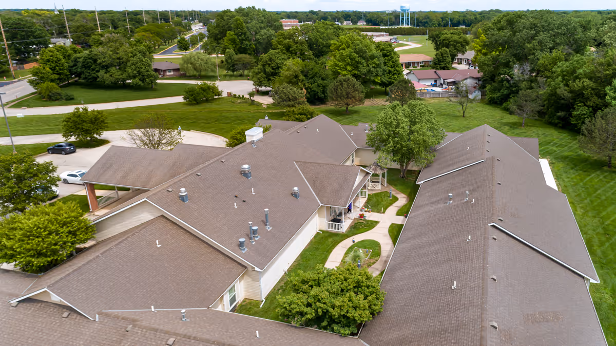 Aerial view of an assisted living facility showing multiple connected rooflines, a central courtyard with walkways, surrounding lawns and trees.