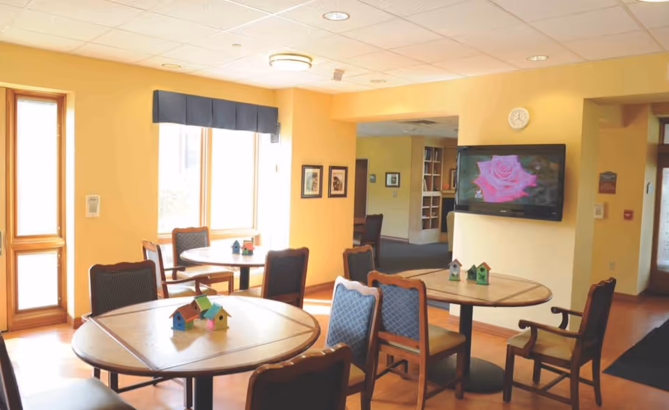 A bright common area in a senior living facility with round wooden tables and cushioned chairs arranged around them. Small decorative birdhouses are placed on the tables. The walls are painted yellow, and there are large windows letting in natural light. A flat-screen TV mounted on the wall displays an image of a pink rose. The room has a warm and inviting atmosphere.