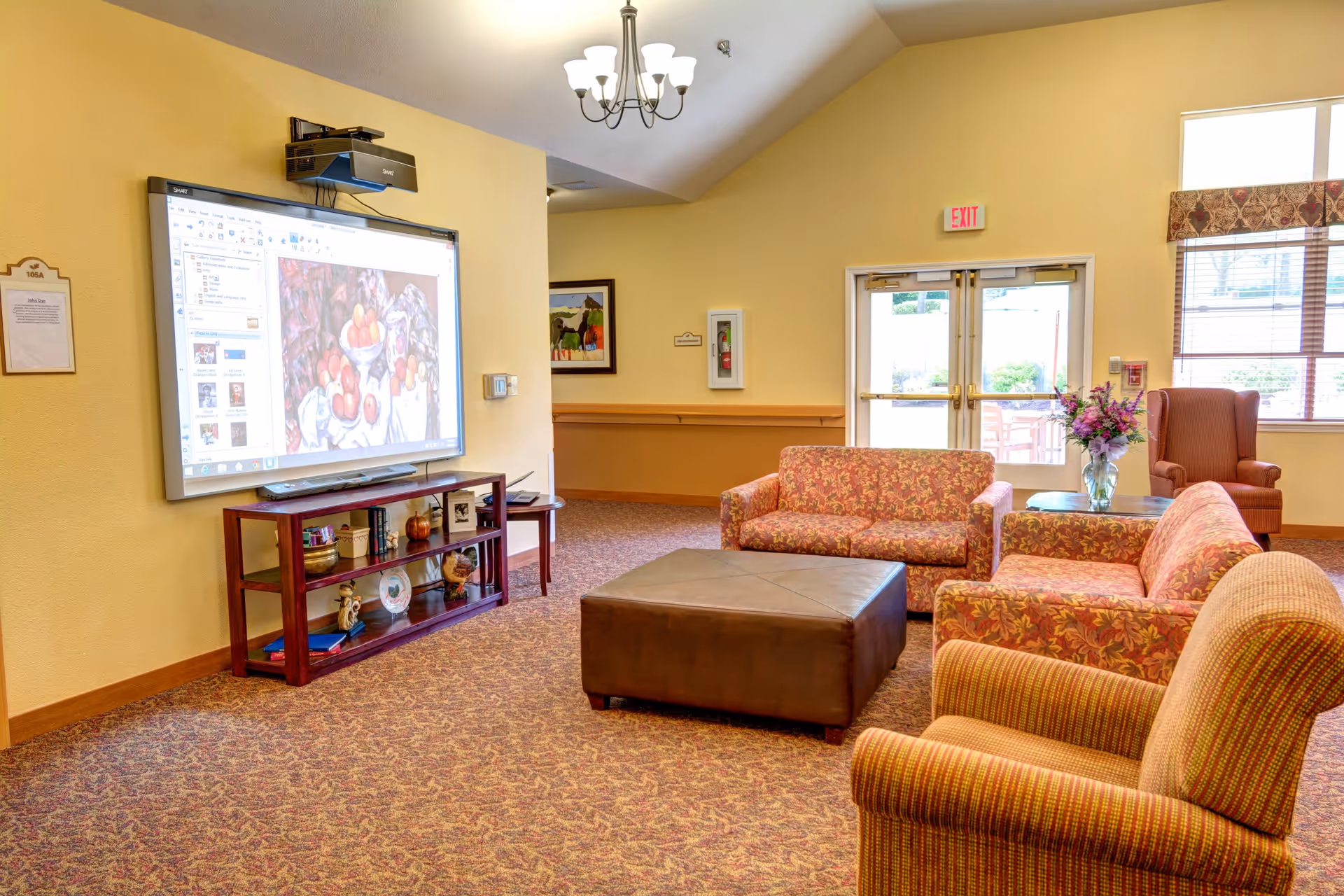 A cozy living room area in Monterey Court Memory Care with patterned sofas and armchairs arranged around a large brown ottoman. A flat-screen projector displays an image on the wall above a wooden shelf holding decorative items. The room has beige walls, carpeted floor, a chandelier, and a glass door leading outside with windows letting in natural light.