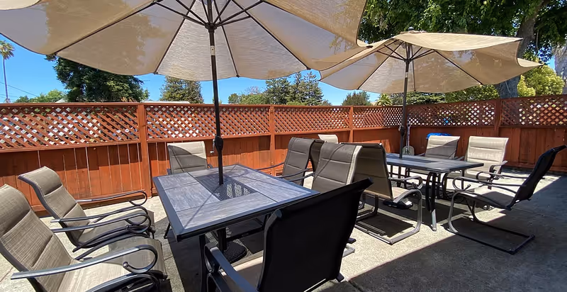 Outdoor patio area with two tables, each surrounded by several cushioned chairs. Large beige umbrellas provide shade over the tables. The area is enclosed by a wooden fence with lattice panels on top, and trees and a clear blue sky are visible in the background.