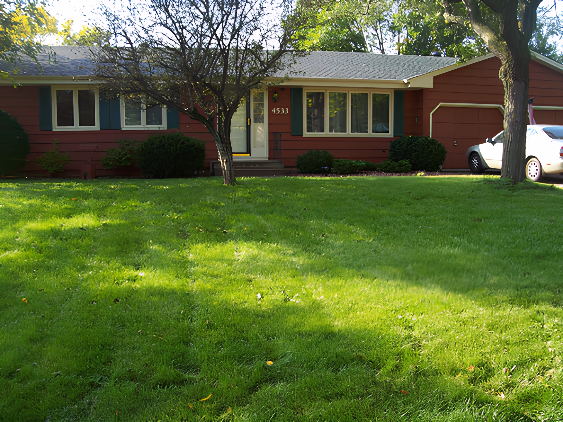 Front view of a single-story red house with a large green lawn, trees, and a white car parked by the garage.