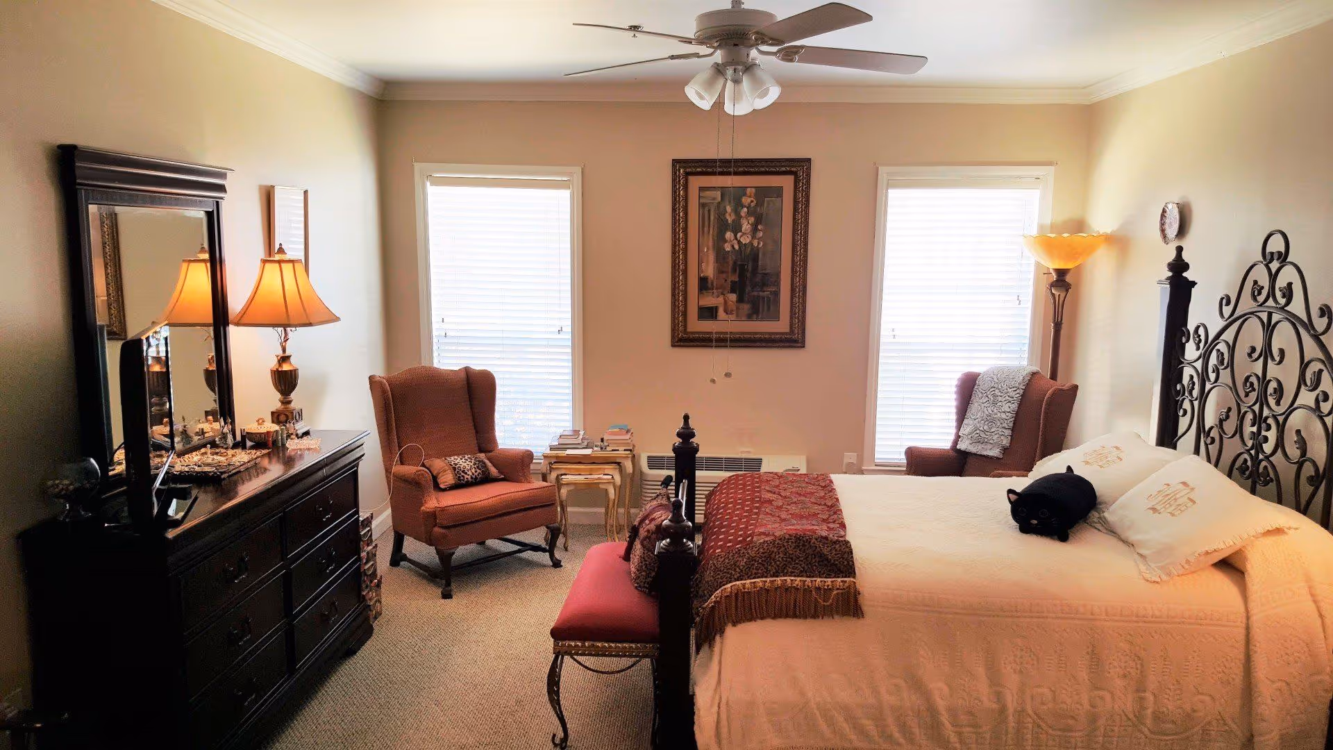 A cozy bedroom with a large bed featuring an ornate metal headboard, white bedding, and decorative pillows including a black cat-shaped pillow. There are two upholstered armchairs near two windows with blinds, a framed painting on the wall between the windows, a dark wooden dresser with a mirror and a table lamp, and a ceiling fan with lights above.