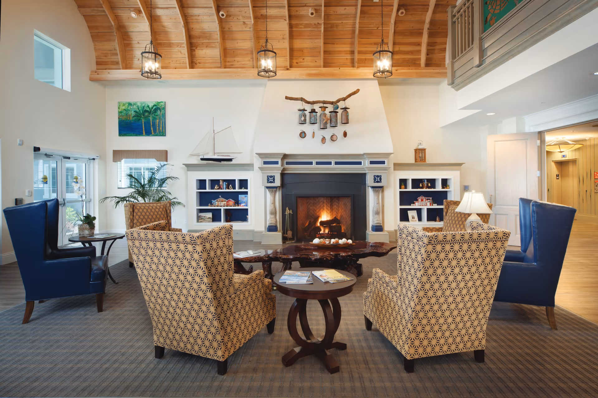 A cozy living room area with a high wooden ceiling and a lit fireplace. The room features four patterned armchairs and two blue leather chairs arranged around a rustic wooden coffee table and a small round side table with magazines. Built-in shelves flank the fireplace, decorated with small models and ornaments. A painting and a potted plant are visible on the left wall, and a hallway with an elevator is seen on the right.