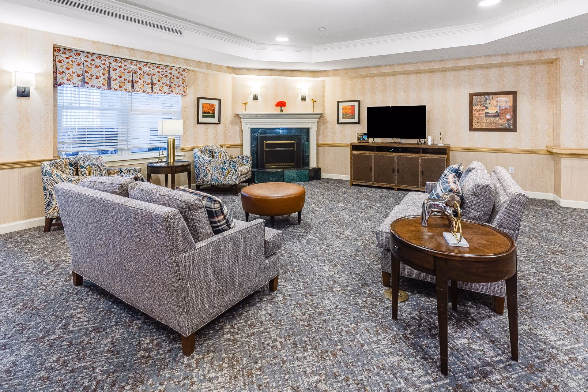 A cozy living room with two gray sofas facing each other, a round brown ottoman in the center, and two patterned armchairs near a window with floral valance curtains. There is a fireplace with a mantel decorated with small items, a flat-screen TV on a wooden cabinet, and framed artwork on the walls. The room has carpeted flooring and soft lighting from wall sconces and a table lamp.