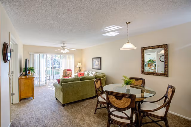 A cozy living and dining area in a senior living facility with a green sectional sofa, a glass-top dining table with four chairs, a ceiling fan, a wall mirror, and sliding glass doors leading to an outdoor balcony.