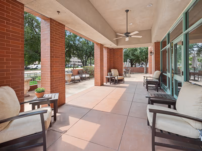 Covered outdoor patio area with cushioned chairs and small tables, brick columns, ceiling fan, and large glass doors leading inside. Trees and greenery are visible outside the patio.