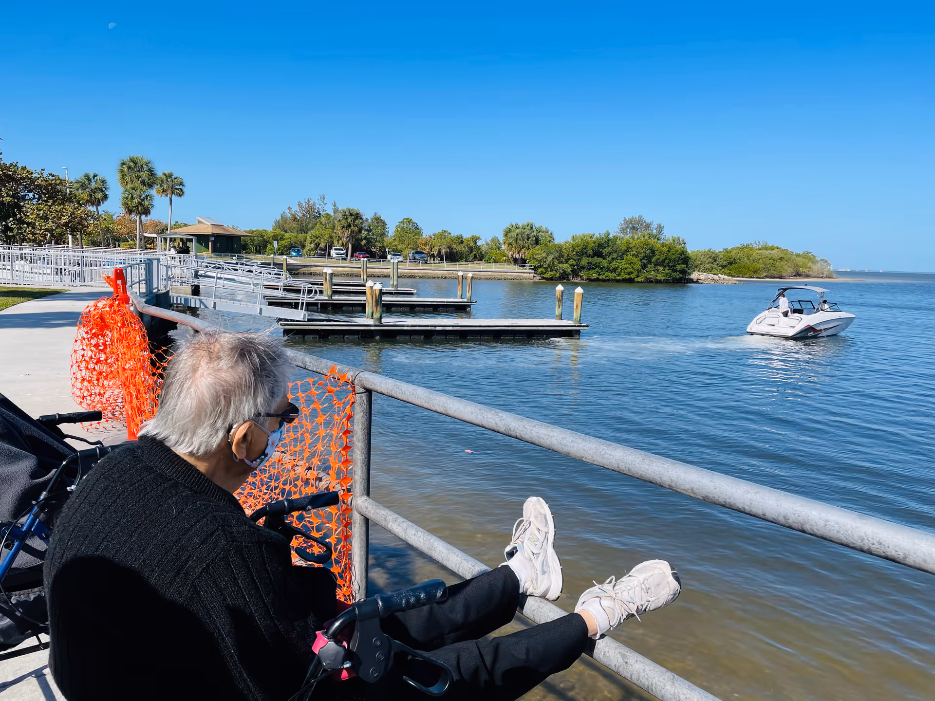 An older adult in a wheelchair sits by a railing at a marina watching a boat on calm water.