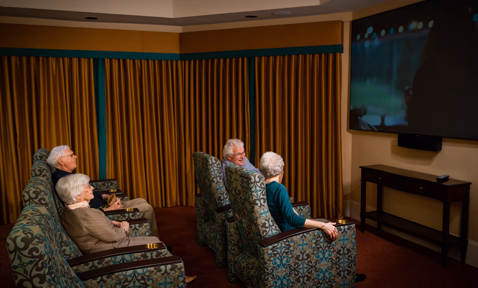 Four elderly residents seated in patterned armchairs watching a movie on a large wall-mounted TV in a dim screening room.
