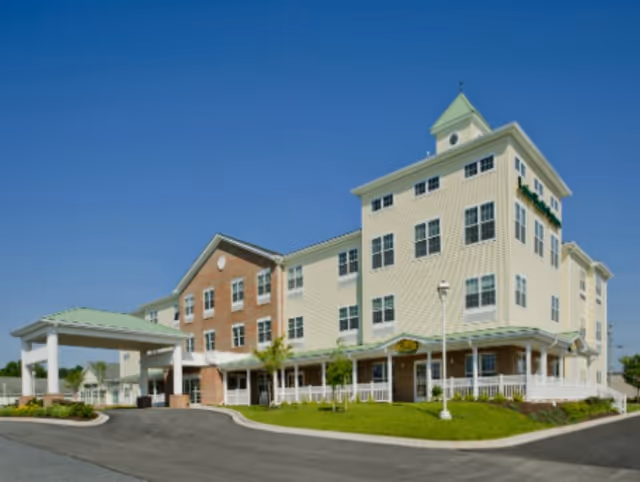Exterior view of a multi-story senior living facility building with a covered entrance, green roof accents, and a well-maintained lawn under a clear blue sky.