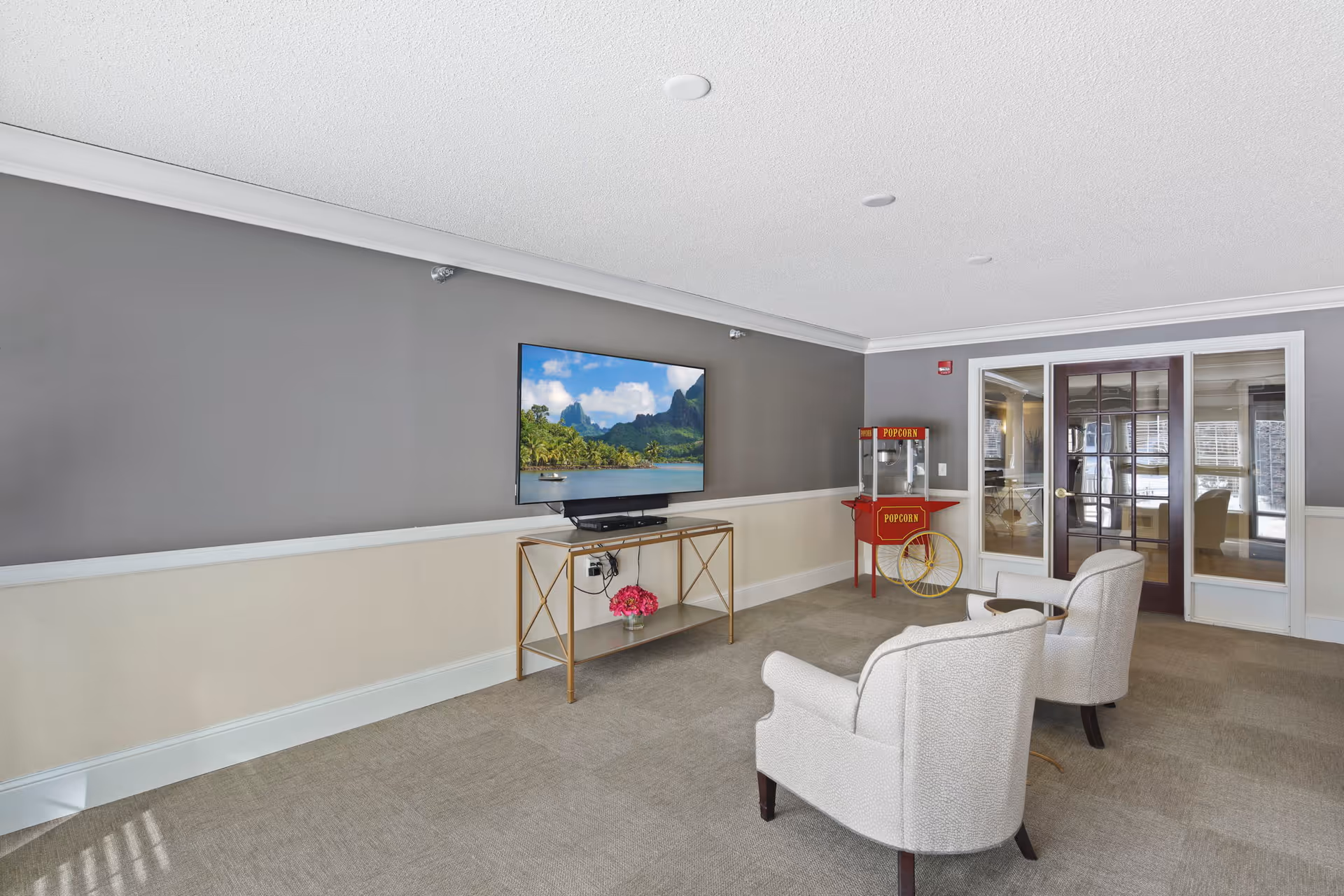 A cozy lounge area with two beige armchairs facing a wall-mounted flat-screen TV displaying a scenic landscape. Below the TV is a gold metal and glass console table with a small pink flower arrangement. To the right, there is a vintage-style red popcorn machine on wheels. The room has gray and beige walls, carpeted floor, and a set of glass double doors leading to another room.