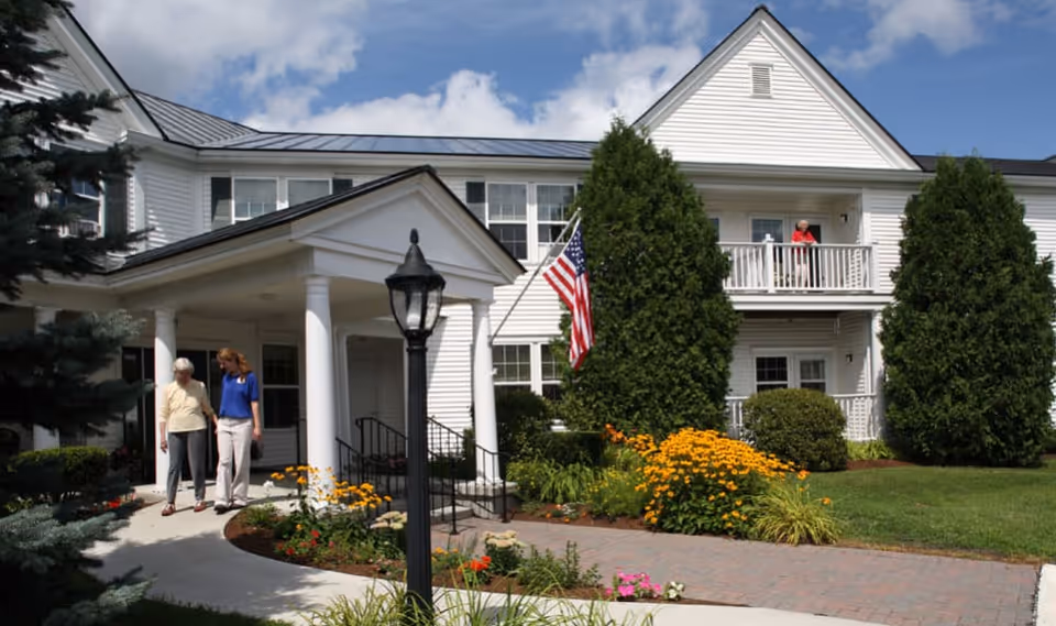 Front exterior of a white senior living building with a covered entrance, American flag, landscaped flowerbeds, and two people walking near the doorway.