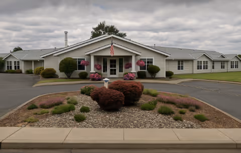 Single-story light-colored assisted living building with a covered entrance, American flag, and landscaped roundabout under a cloudy sky.