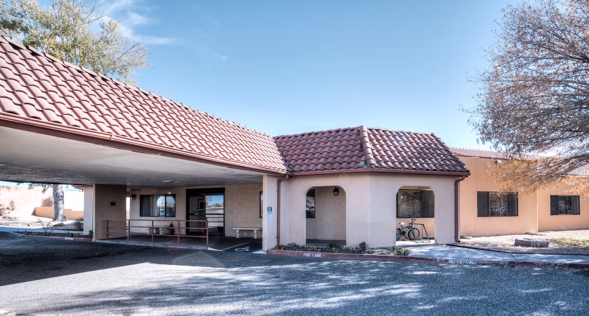 Exterior view of Skies Healthcare and Rehabilitation Center showing a single-story building with a red tiled roof, a covered entrance with a ramp, a bicycle parked near a window, and a tree with sparse leaves on the right side under a clear blue sky.
